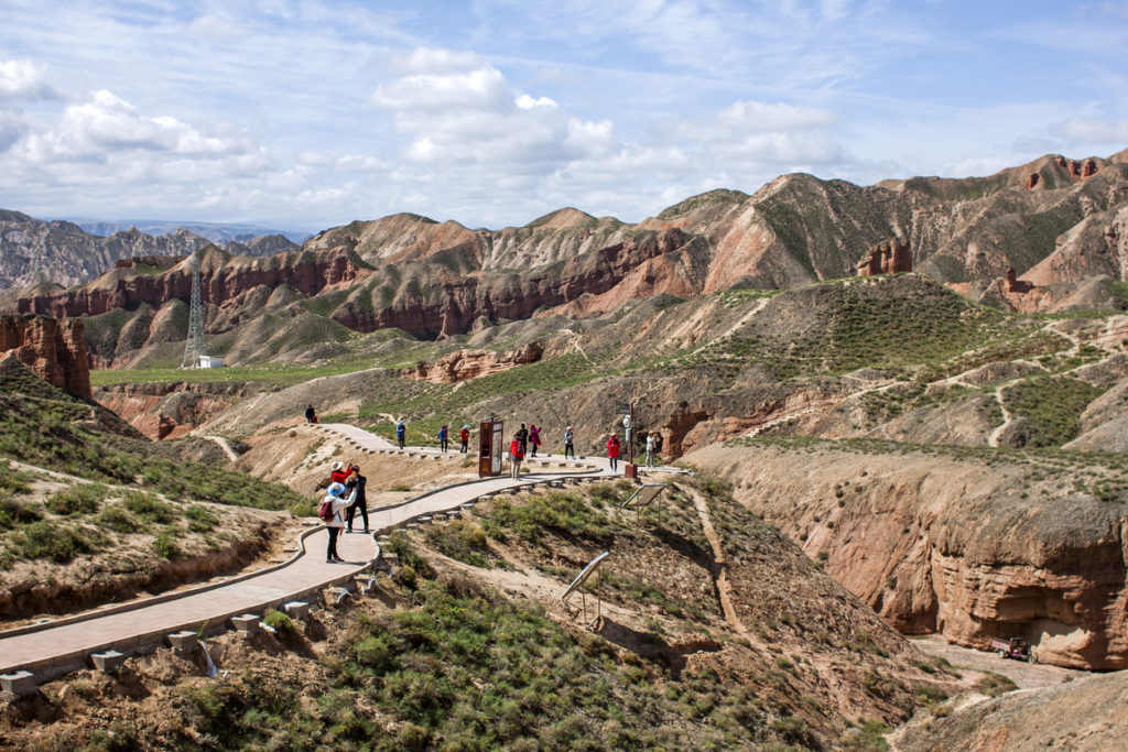 Group of Chinese Tourists are Walking Along a Path in Binggou Danxia Canyon Landform in Zhangye, Sunan Region, Gansu Province, China. Red Sandstone Rocks in the Geopark. Clouds and Blue Sky on a Sunny Day.